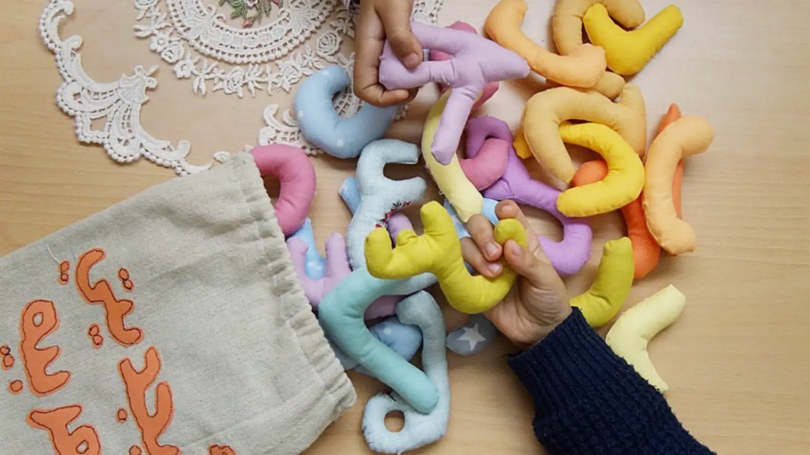 Soft fabric Arabic letters in pastel colours spilling from a cloth bag, with a child's hands playing among them.