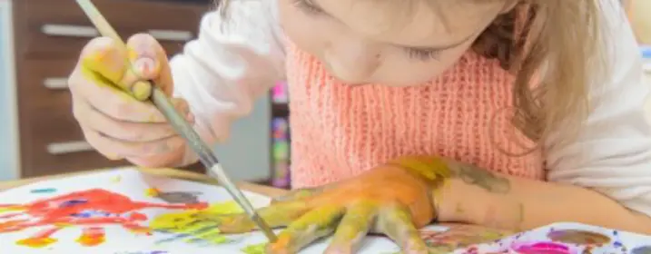 طفلة ترسم بالفرشاة على يدها الملونة فوق ورقة.